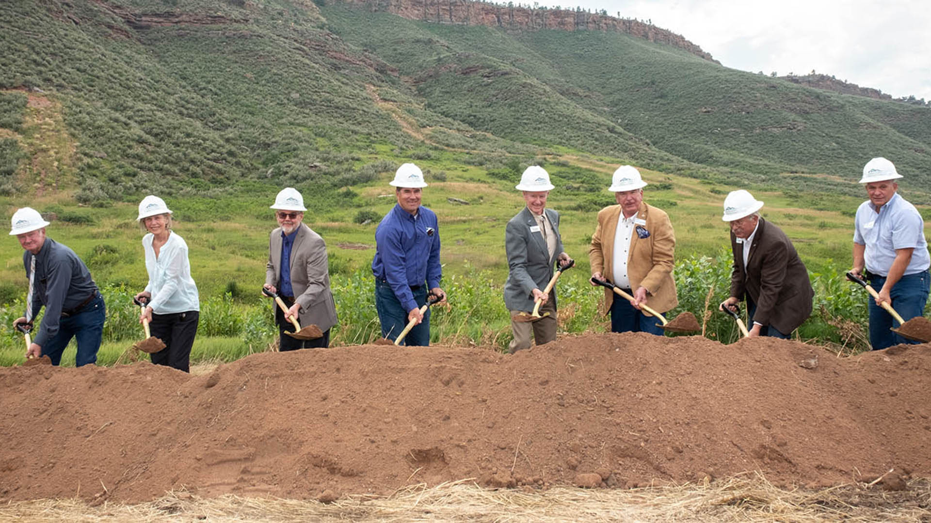 Eight people in hardhats holding gold shovels in front of a pile of dirt. 
