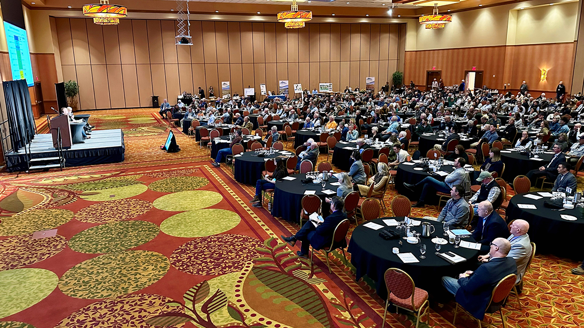 A banquet room full of people sitting at tables watching a panel present on stage
