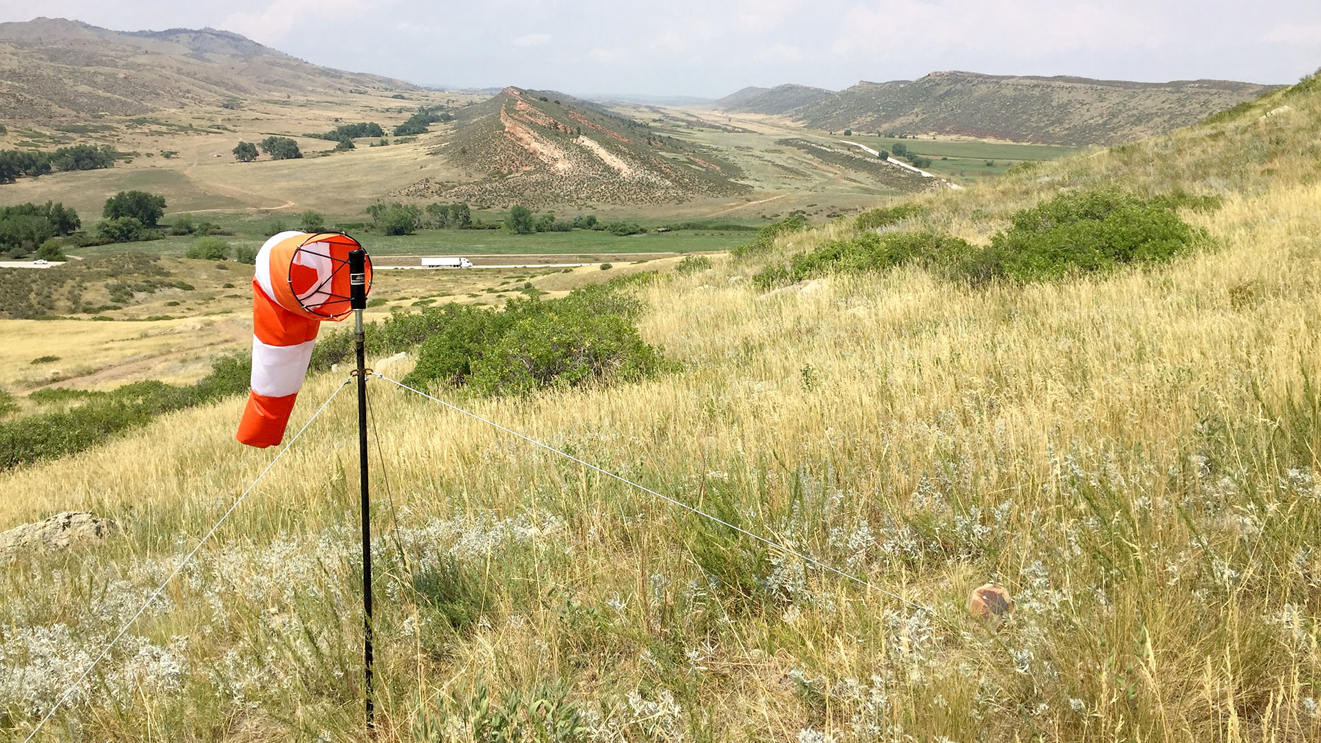 Orange and white windsock flying over some grassland with a highway below in the valley.
