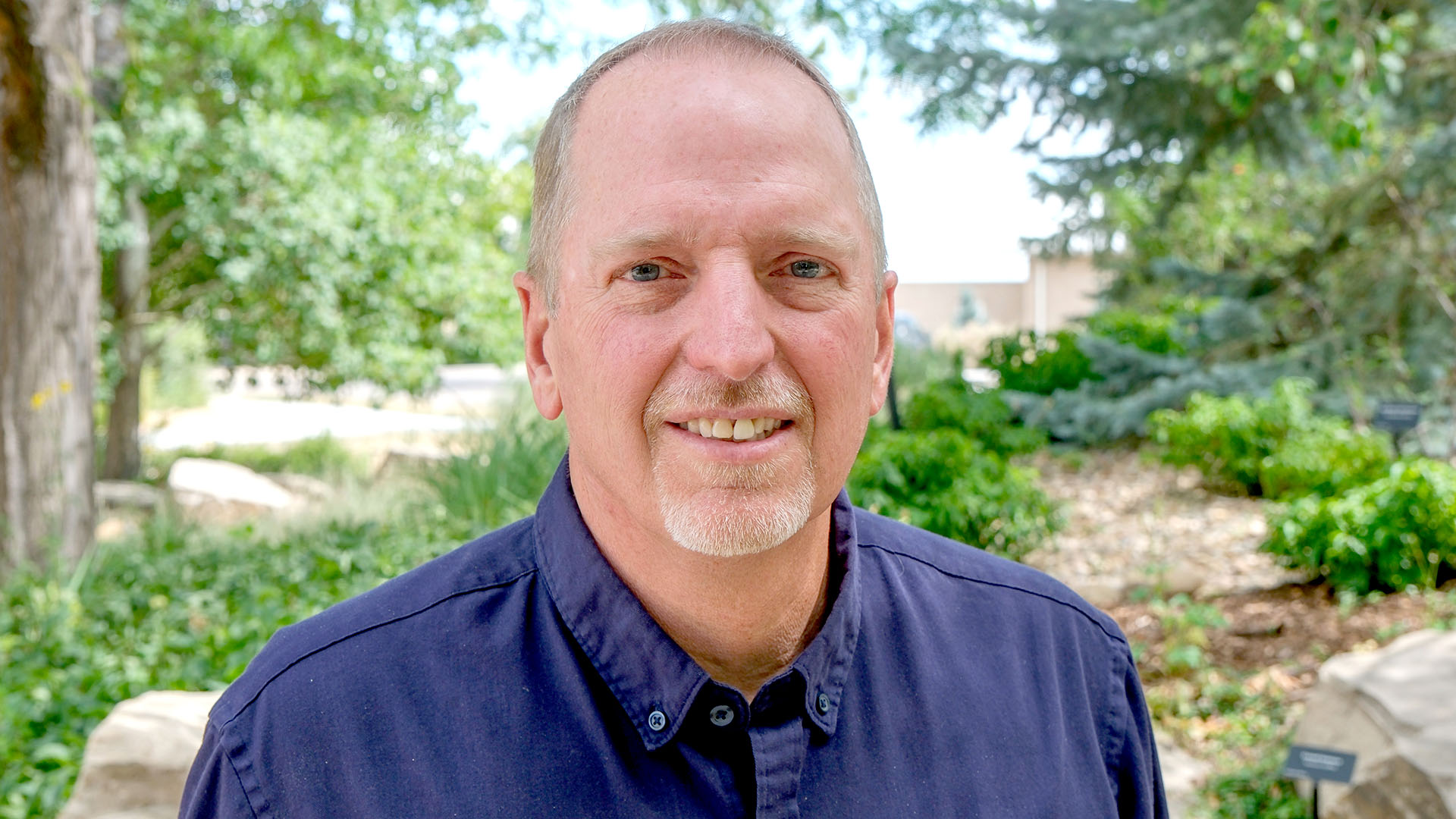 Man wearing a blue button up shirt standing with trees and plants behind him. 