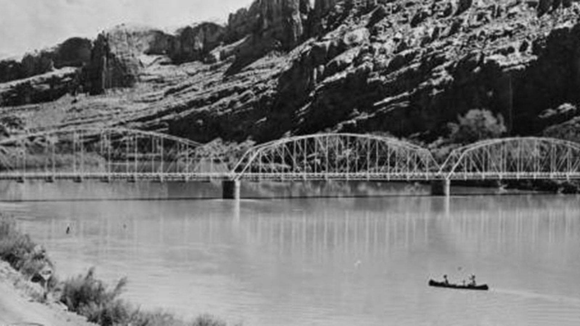 Historic image of a bridge over the top of the Colorado River with a canoe and 2 people in it.