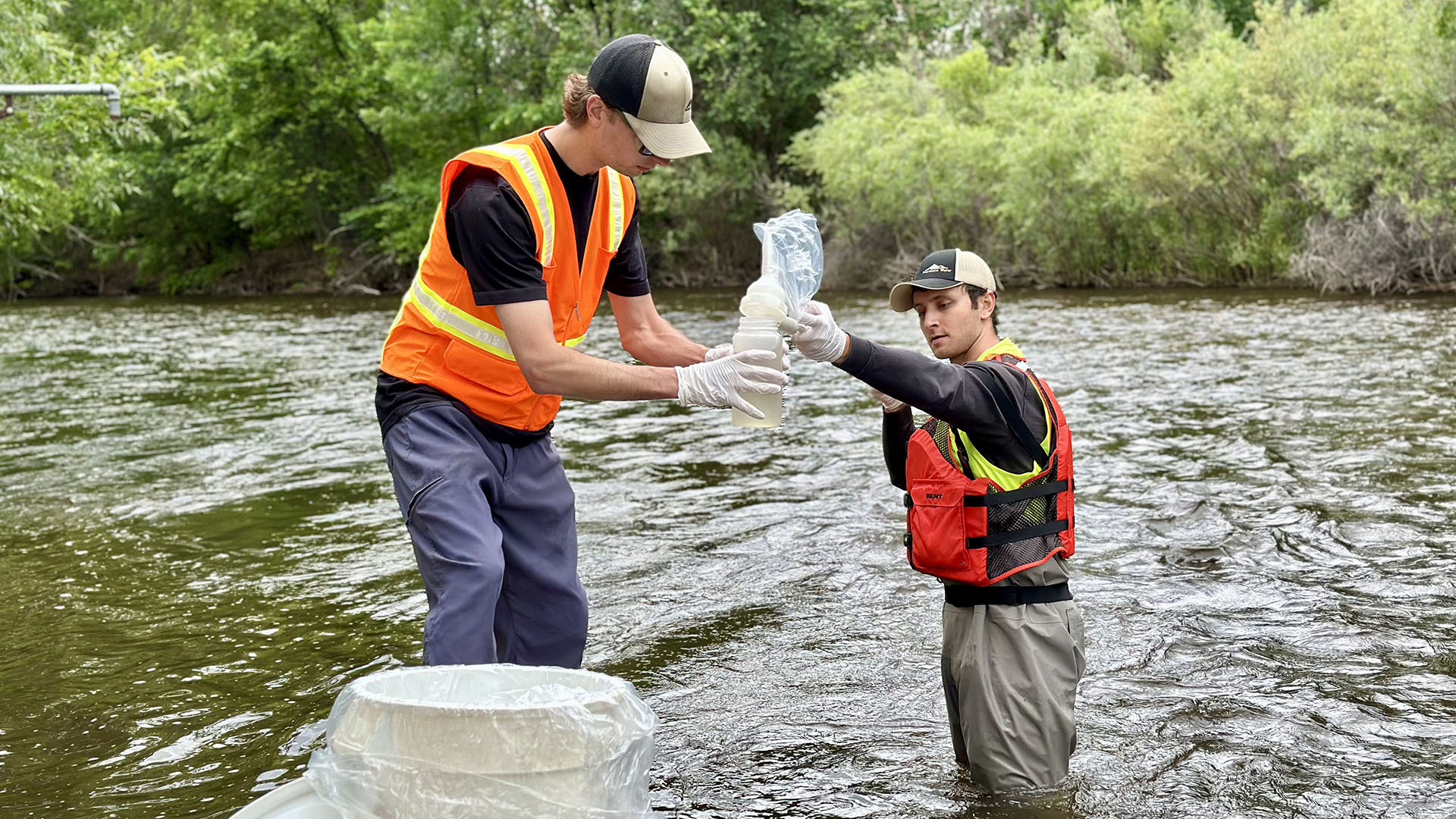 Two men wearing life vests in a river collecting water quality samples.