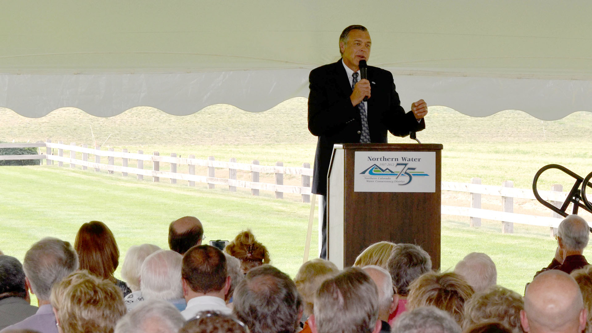 Man at a podium under a tent addressing a crowd.