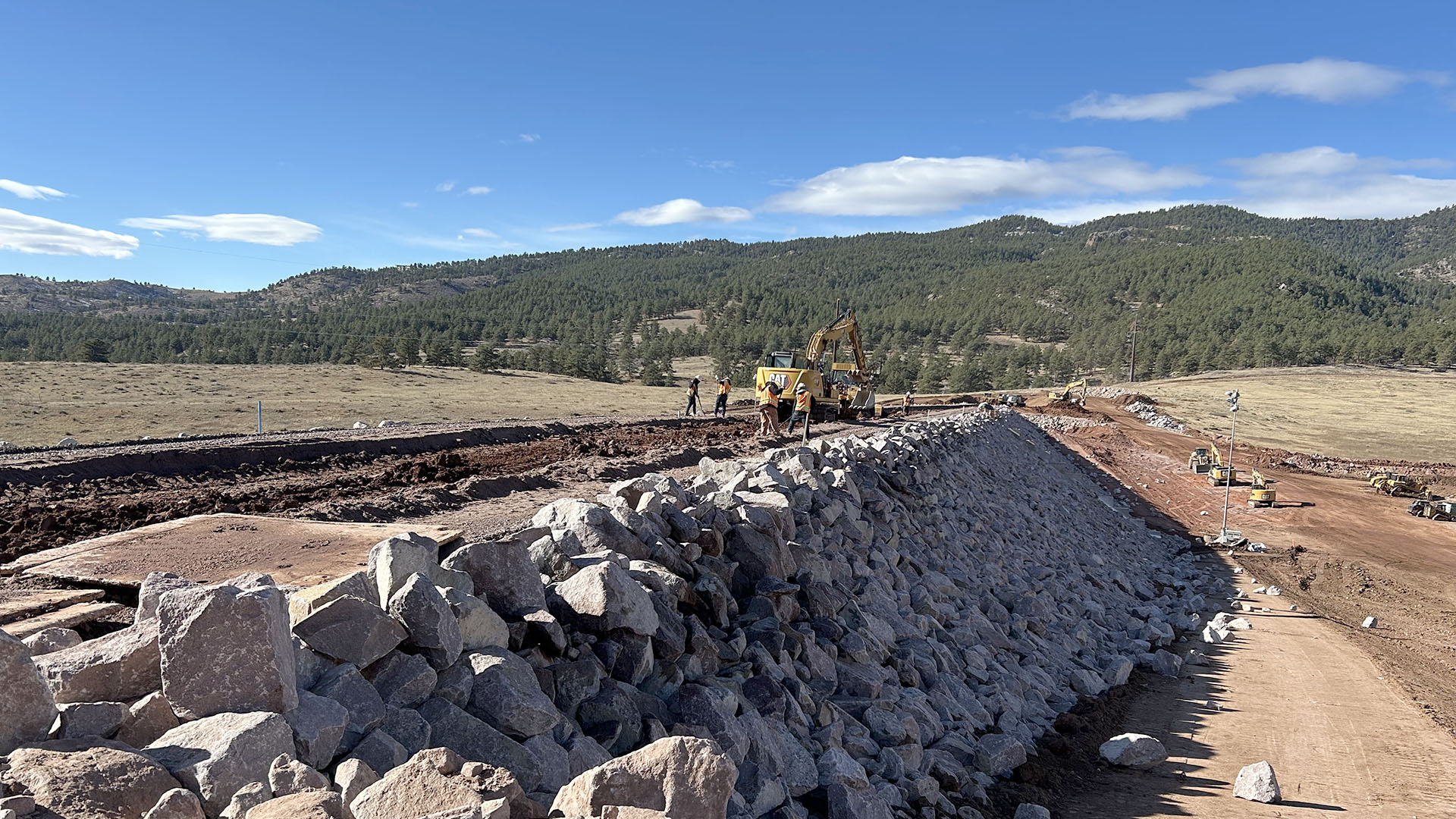 Backhoe and people standing on top of a dam with rocks on side. 
