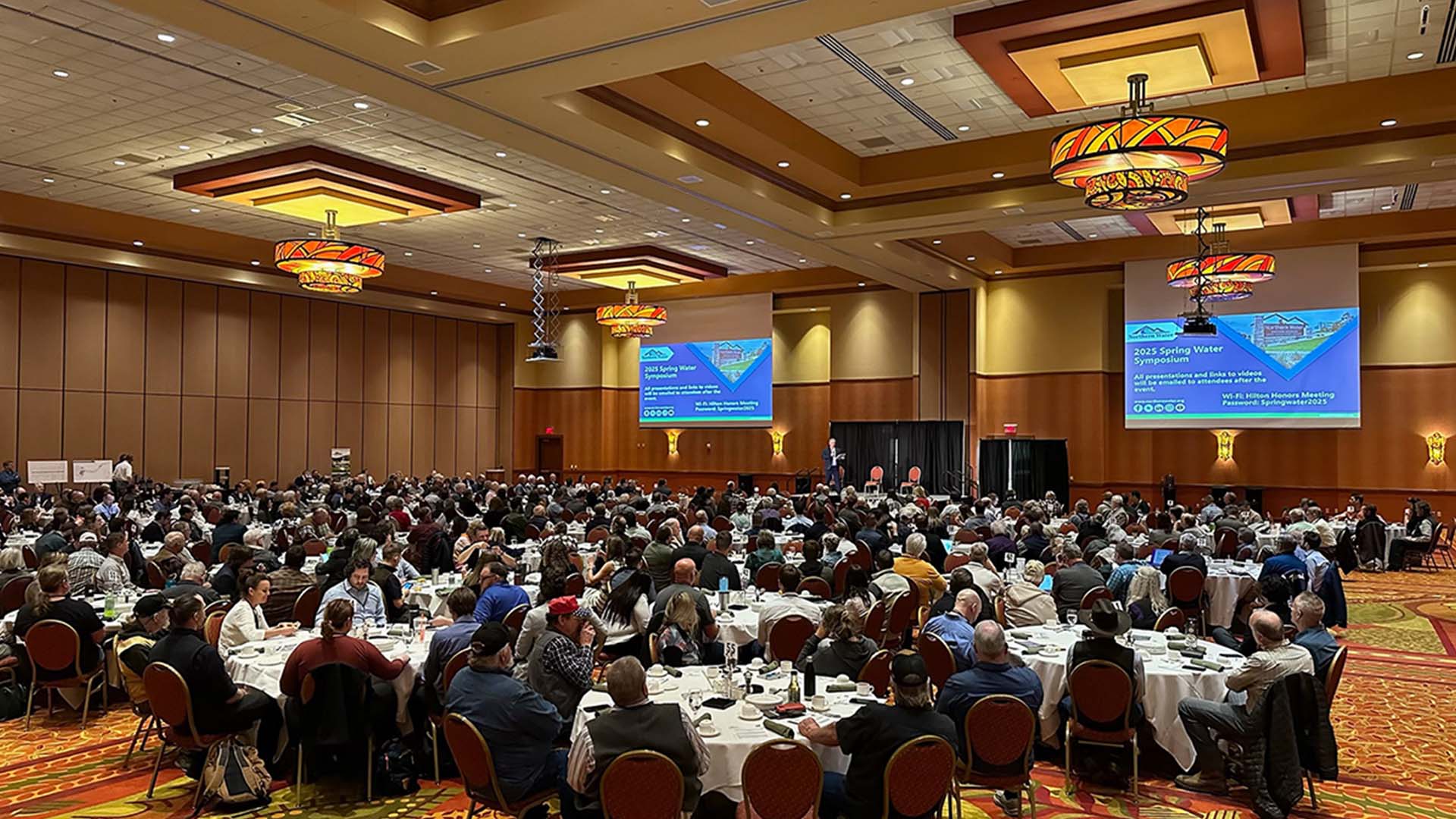 A room full of people at a Symposium with large screens in front and overhead lights.