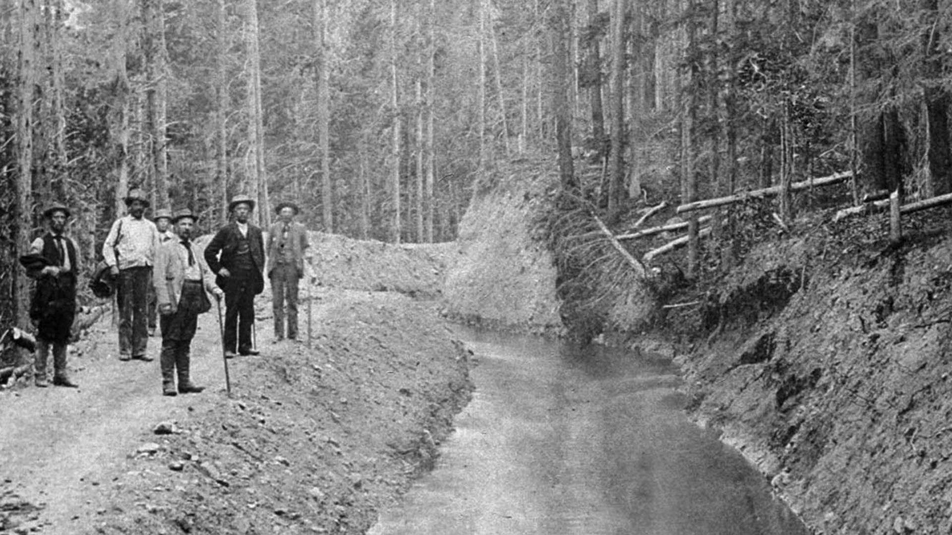 Historic image of 6 men in suits and top hats next to an irrigation ditch. 