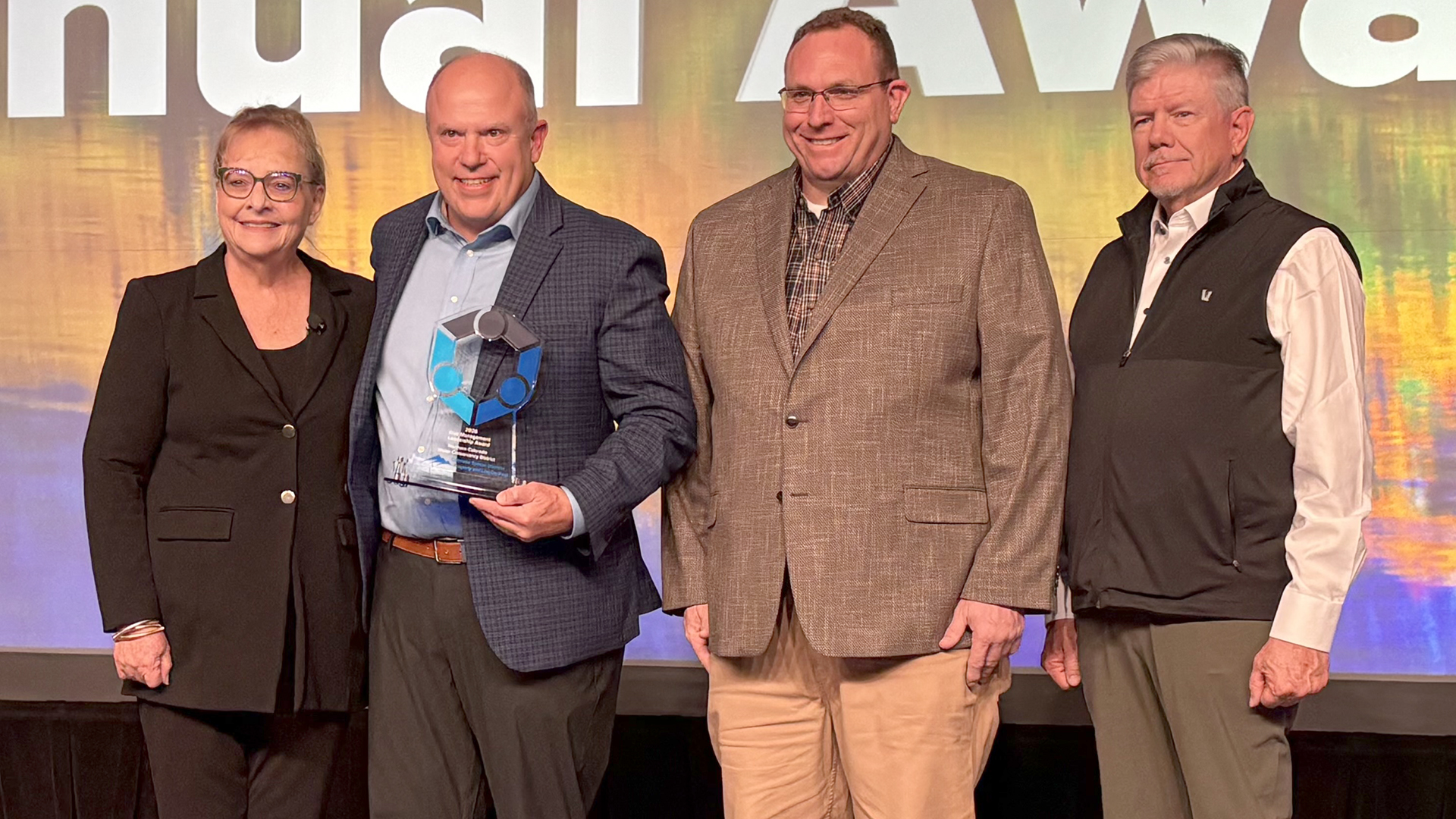 A lady presenting a glass award to three men. Screen behind them with images. 