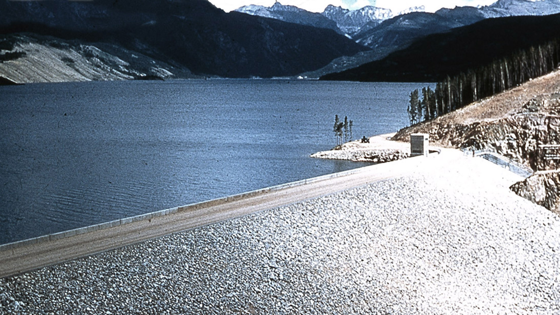 Dam in front of image with water held behind and mountains in background.