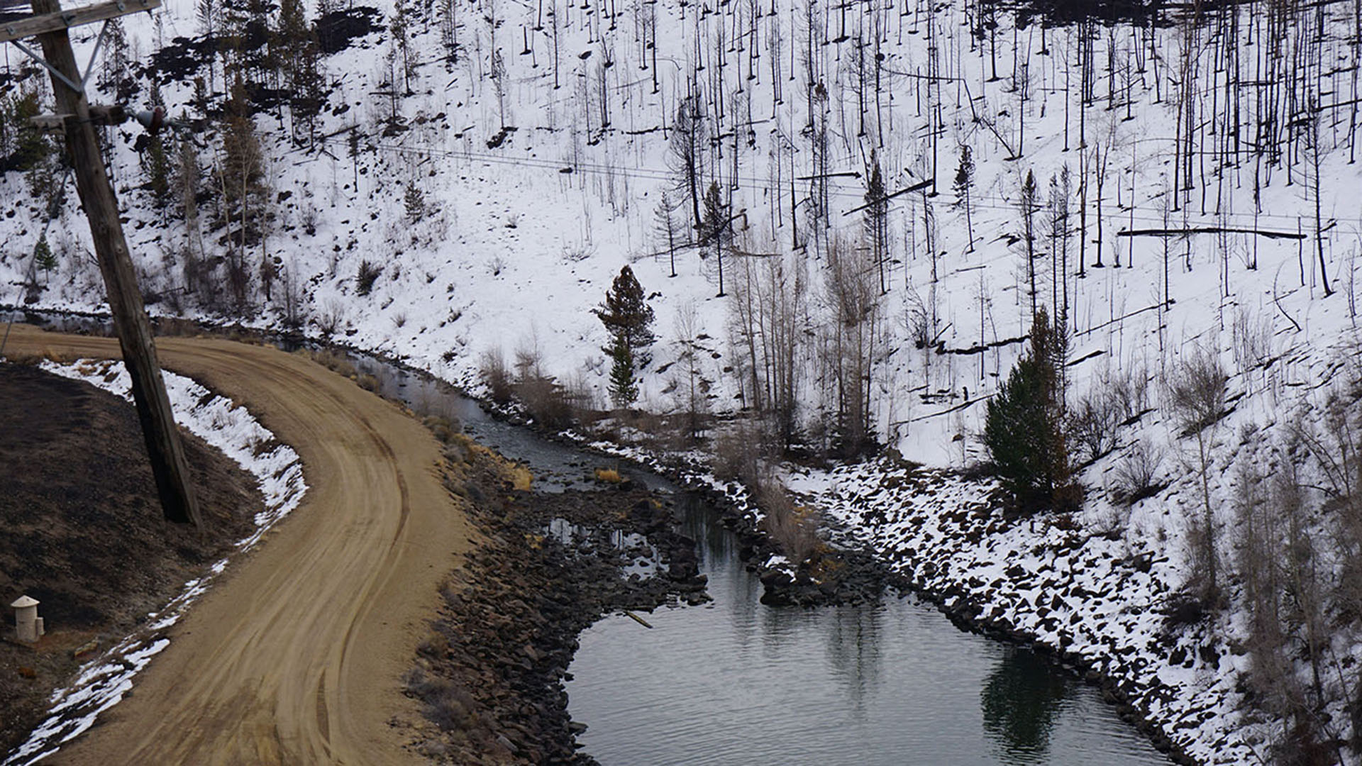 A creek with burned trees on the hillsides next to it with a dusting of snow.