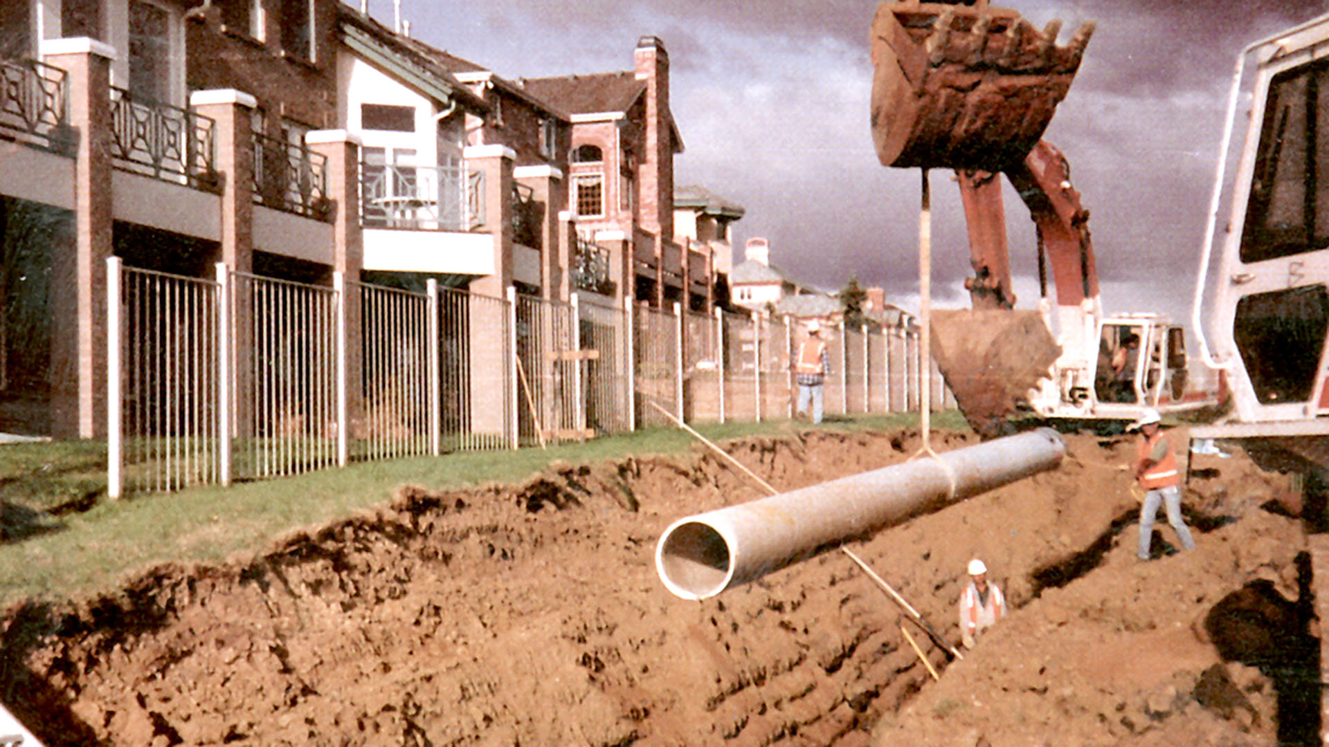 A backhoe lifting a pipe into the ground with homes behind it.