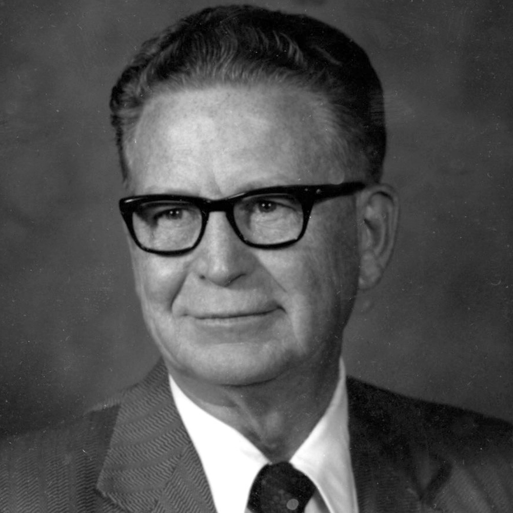 Grayscale image of man wearing a suit with black thick glasses.