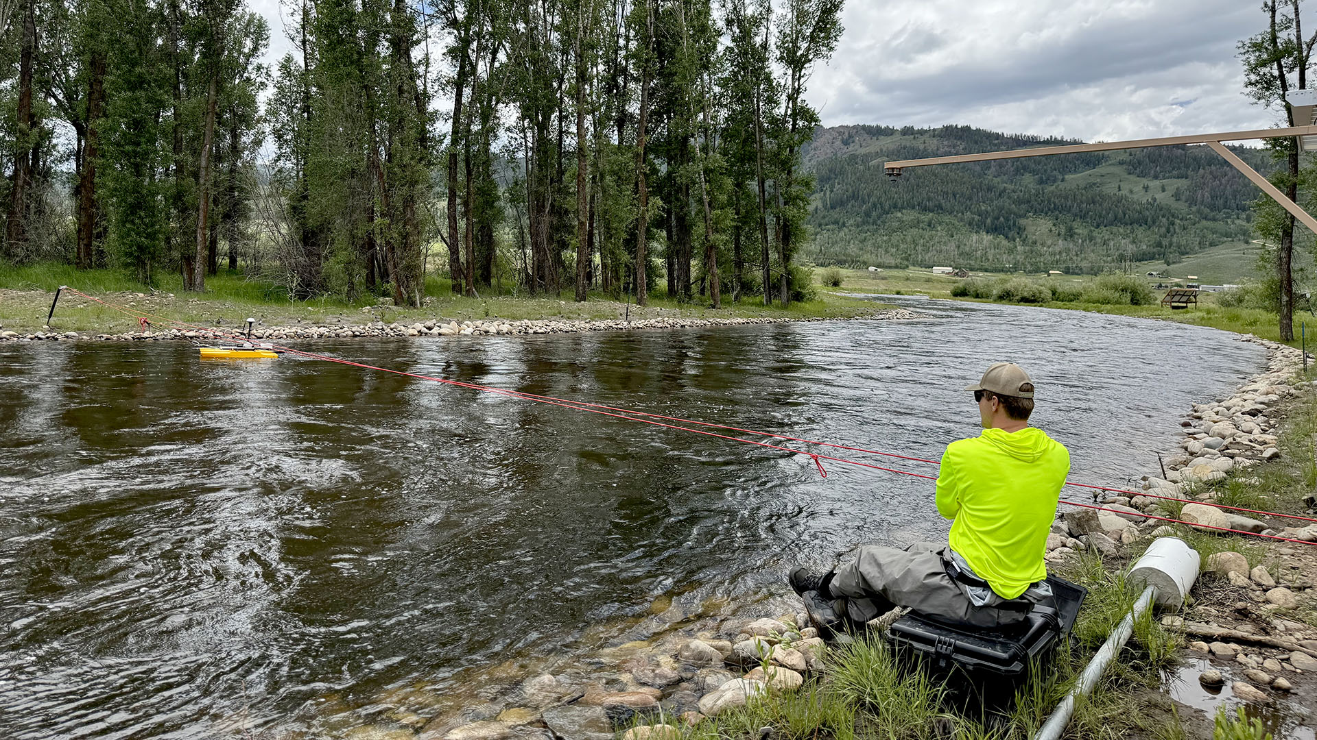 Man in bright yellow shirt sitting next to river conducting flow measurements.