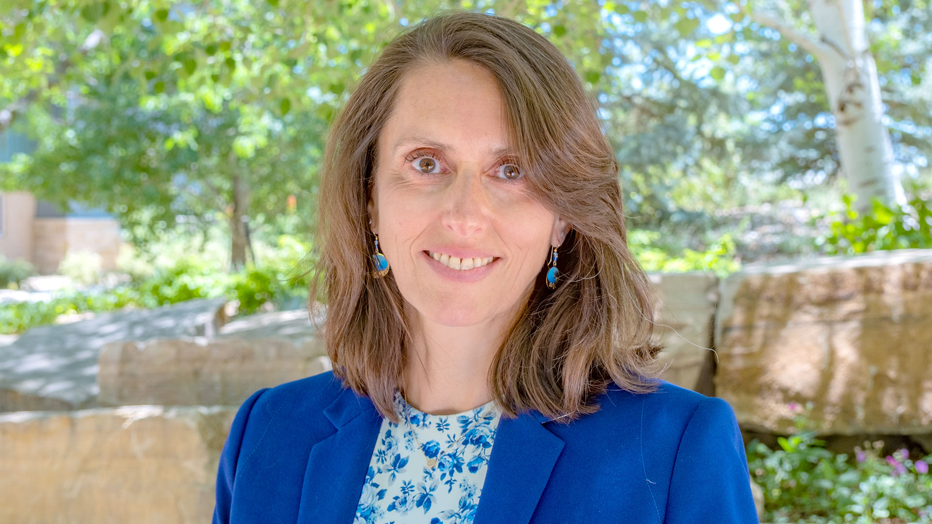 Woman in a blue suit with print shirt underneath and shoulder length hair.