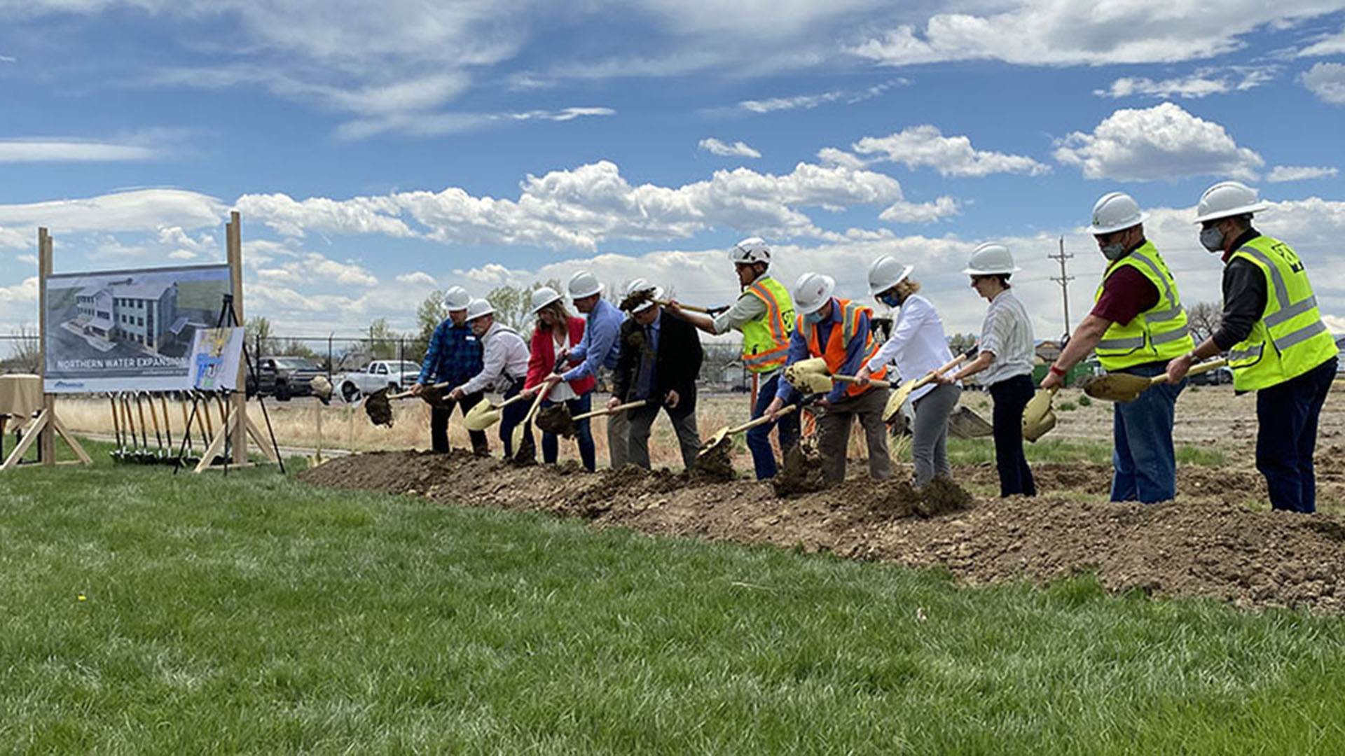People lined up wearing hardhats with gold shovels turning dirt for a groundbreaking event.