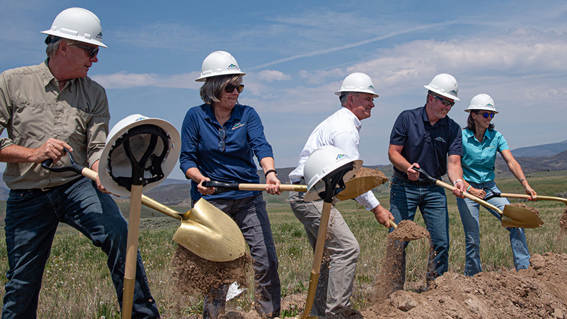 Five people wearing hardhats holding gold shovels turning over dirt. 