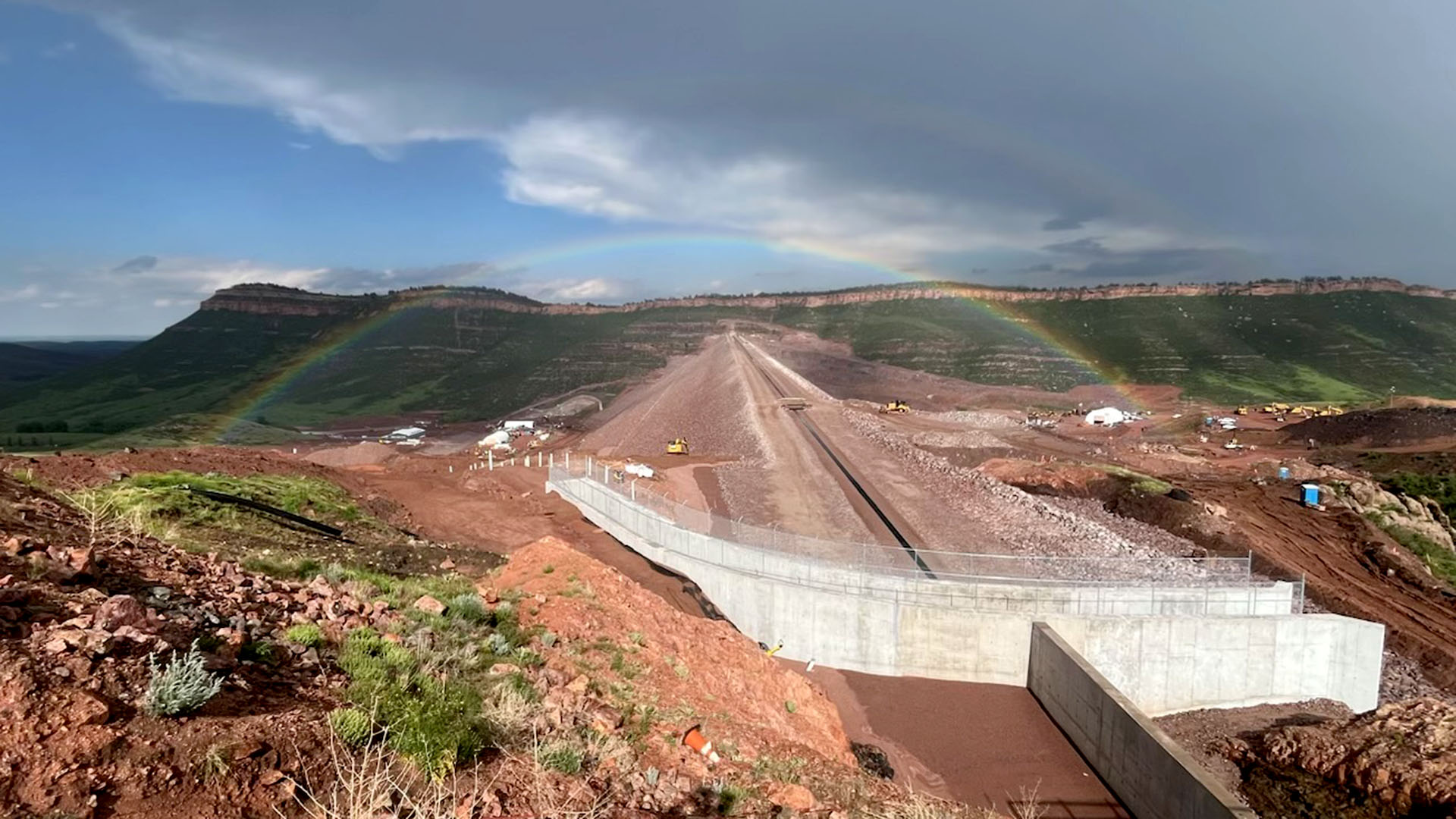 Main dam at Chimney Hollow with concrete spillway in foreground and rainbow in sky above.