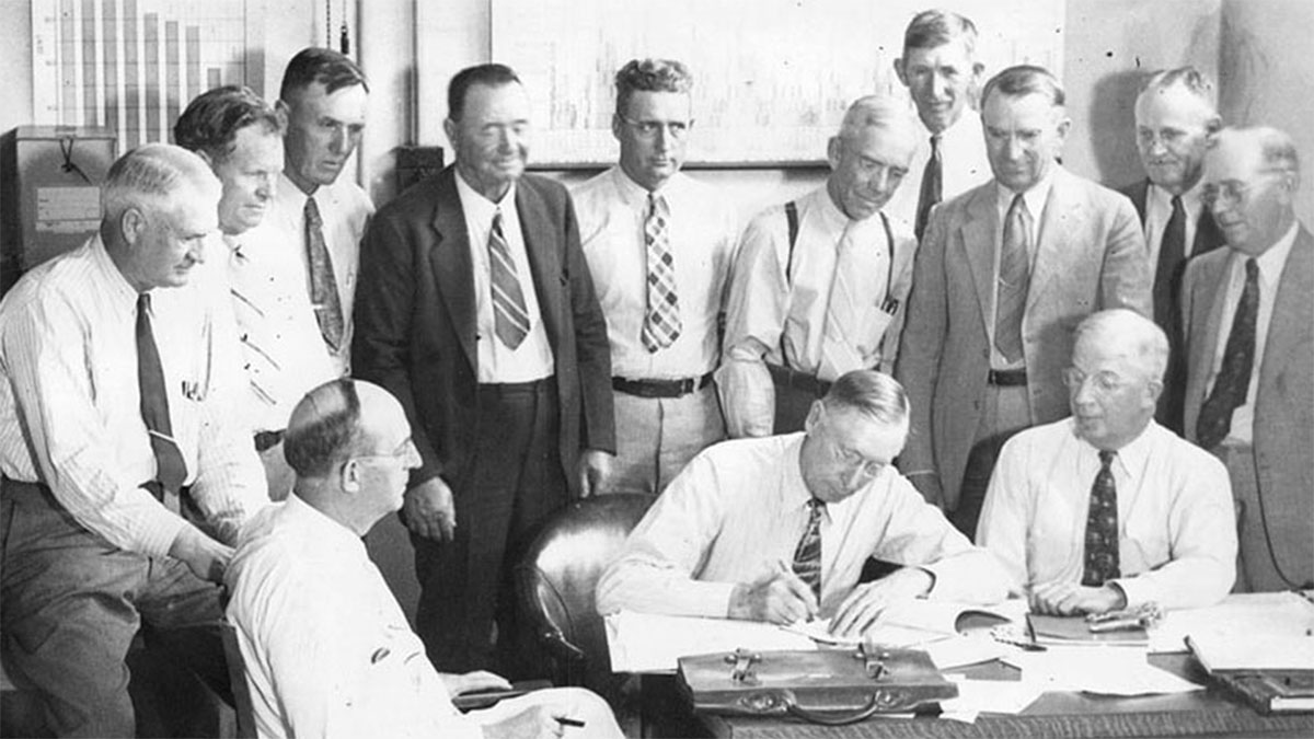 Grayscale photo from 1937 shows a group of male legislators, three sitting and one signing the Repayment Contract.