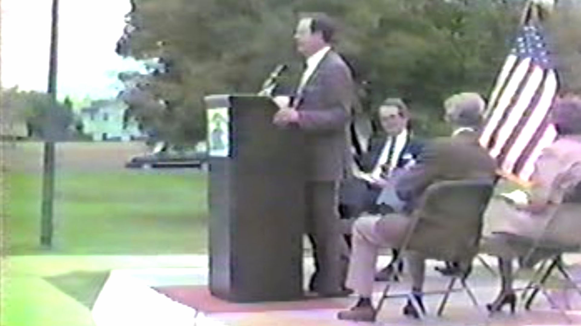 Man standing at a podium with other people on chairs behind him. 
