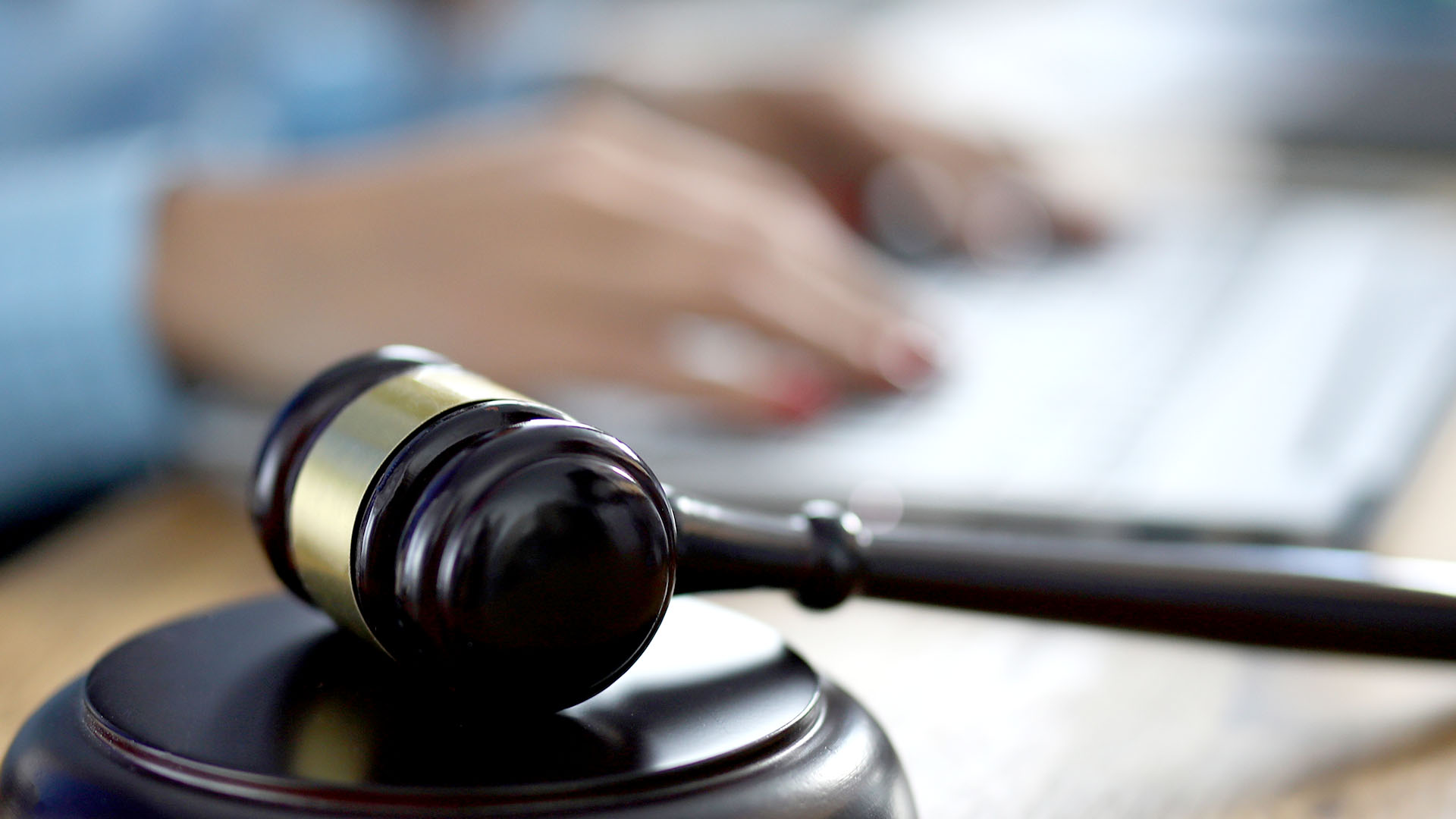 Gavel on a desk in the forefront with a hand on papers in the background.