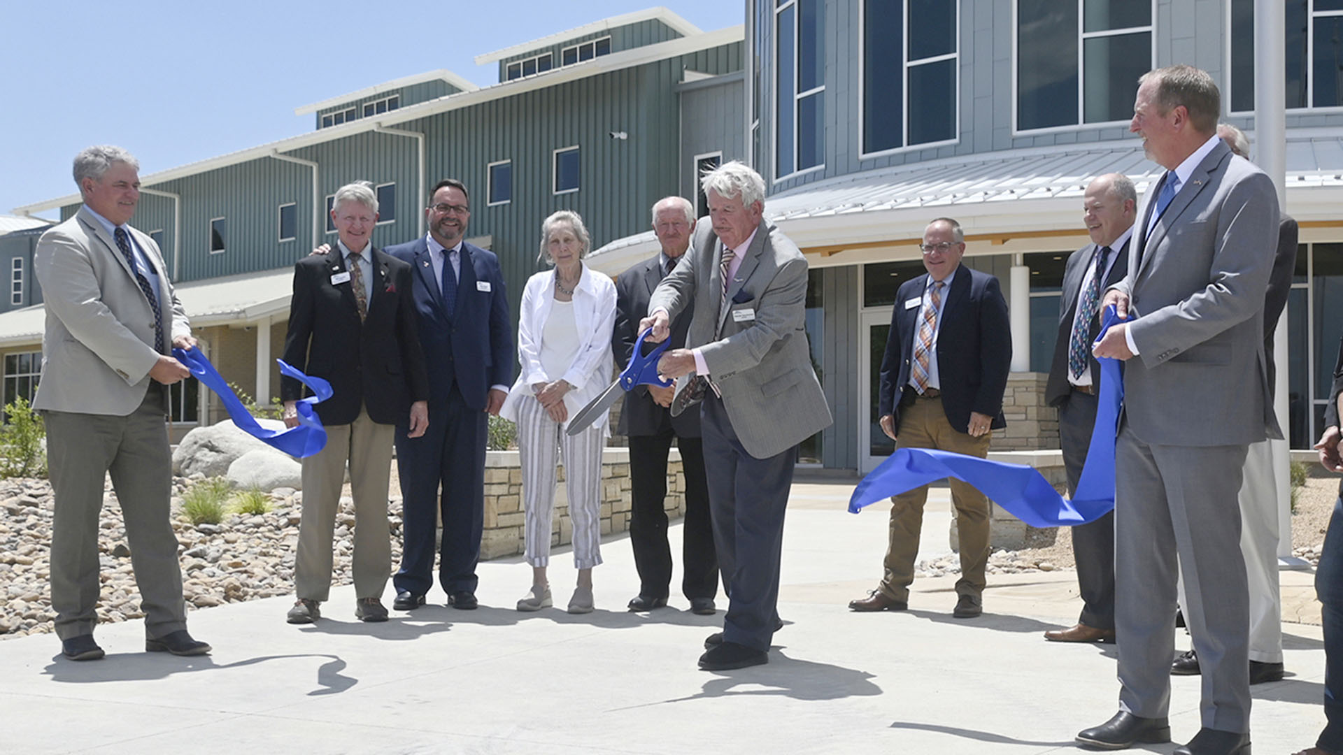 People lined up in front of a building holding a large blue ribbon to cut.