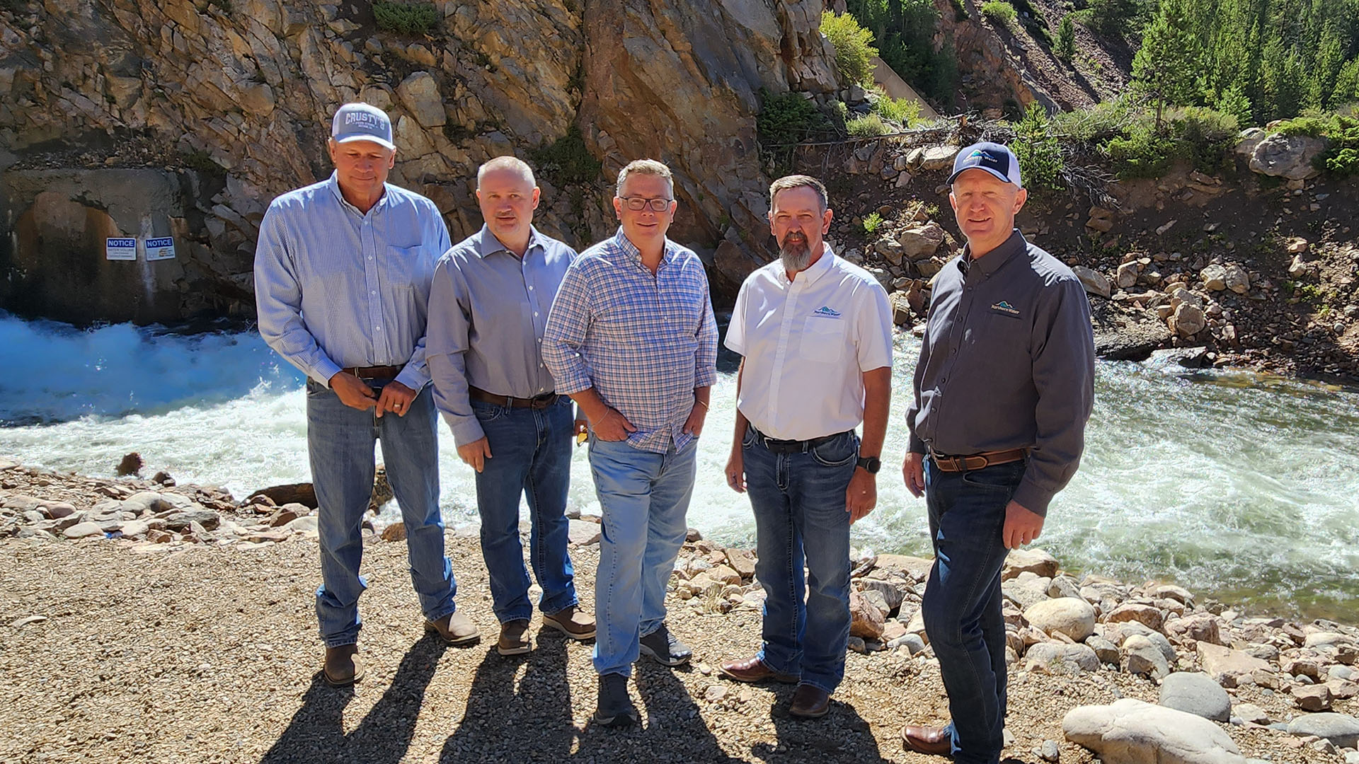 Five men stand in front of a flowing river with a rocky brackdrop