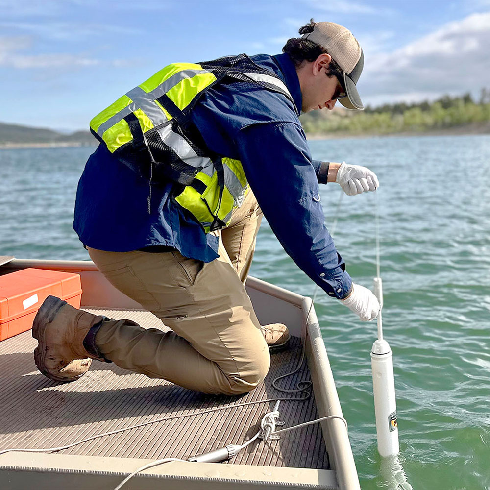 Man in a life vest on a boat dropping a water quality monitoring tool into the water.