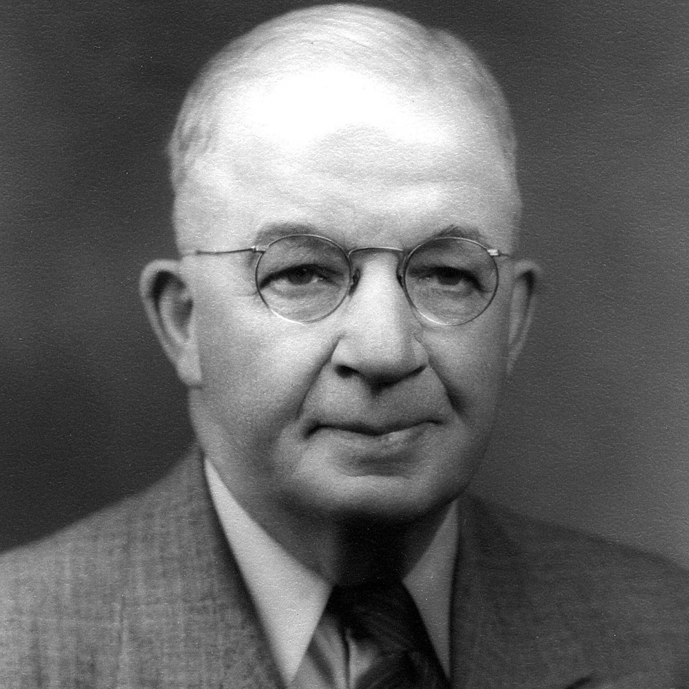Grayscale image of a man in a suit wearing wire framed glasses.