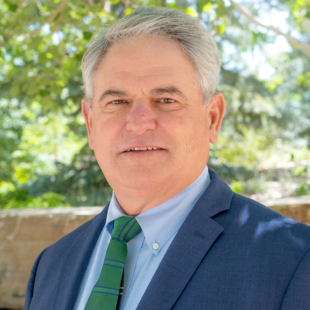 Man in a blue suit with green tie outside with trees behind.