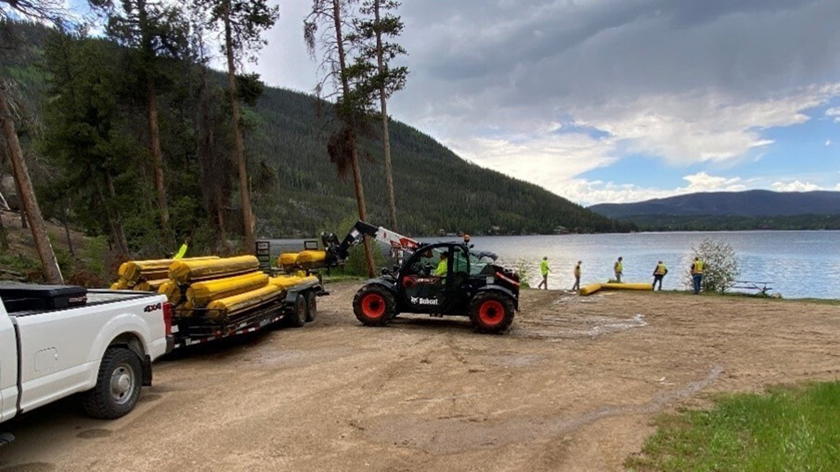 Yellow debris booms are being removed from a lake and placed onto a trailer pulled by a white pickup truck.