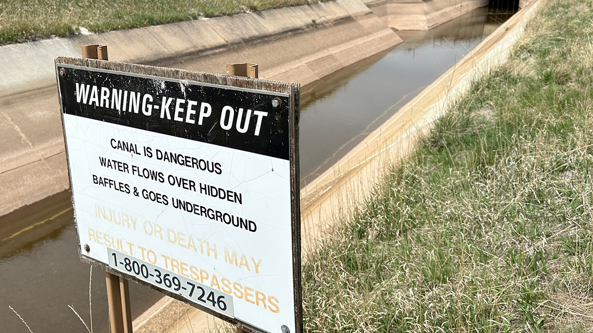 A sign in front of a canal says "warning, keep out." In back on the left side of the canal is a ramp that game animals can use to escape.